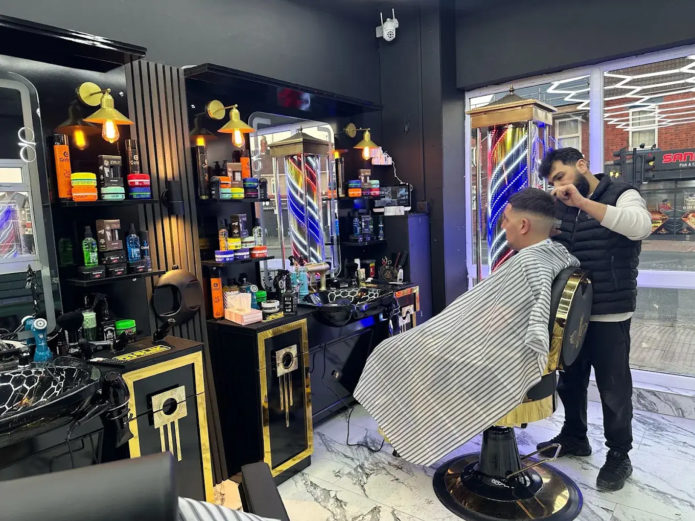A barber cutting a client in a striped cape at a black and gold station, the street outside the window, at HAWAR BARBER in Darlington