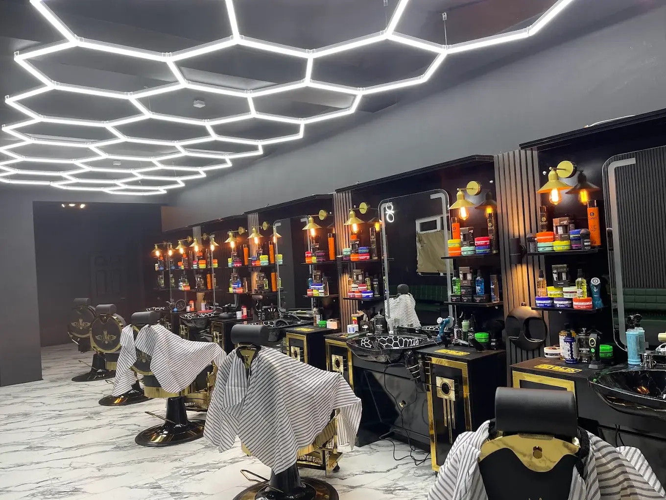A row of black and gold chairs with striped capes ready under the hexagon ceiling lights at HAWAR BARBER in Darlington